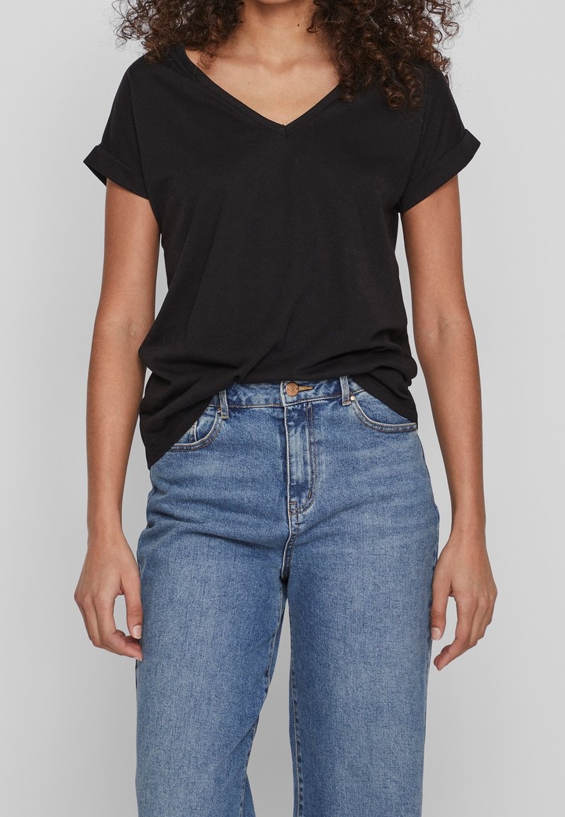 T-shirt noir à col en V avec manches retroussées, associé à un jean bleu taille haute. Le tissu semble doux et décontracté, offrant une coupe ample et confortable.