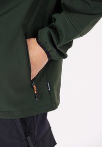 Veste verte en tissu lisse avec des poignets élastiques. Comprend une poche latérale zippée avec un tirant orange et des accents noirs.