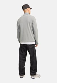 Chaqueta de forro polar en gris claro con cuello alto, combinada con unos vaqueros de mezclilla oscura y zapatillas de color claro. Vista trasera con diseño simple.