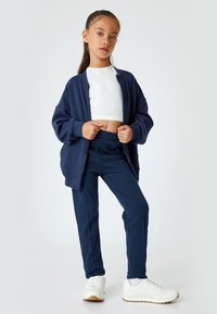 Granatowa kurtka na zamek błyskawiczny, biały crop top i dopasowane granatowe spodnie. Ubrane białe sneakersy z teksturowaną podeszwą. Prosty, czysty design.
