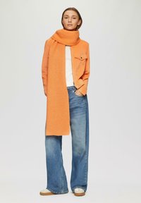 Oranje gebreide sjaal over een lichtoranje corduroy jasje, gecombineerd met wijde blauwe jeans en witte sneakers. Eenvoudige, textuurrijke, casual uitstraling.