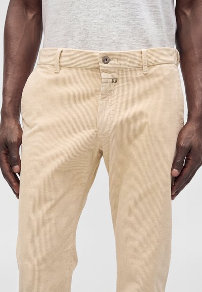 Homme portant un pantalon en velours côtelé beige et une chemise texturée gris clair, les mains détendues le long du corps devant un fond uni.