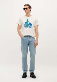 Tricou crem cu grafică de munte albastră, blugi decolorati, și pantofi negri. Modelul poartă ochelari de soare negri și stă în fața unui fundal neutru.