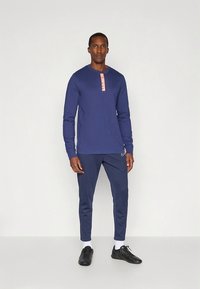 Tengerészkék hosszú ujjú ing rózsaszín gombos előlappal és logóval, tengerészkék karcsúsított nadrággal és fekete sportcipővel párosítva.