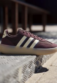 Baskets bordeaux avec des bandes blanches Adidas et une semelle translucide reposant sur une marche en métal texturée à l'extérieur au soleil.