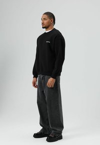 Sweat-shirt noir à col ras du cou avec logo brodé, assorti à un jean gris foncé ample et des chaussures noires épaisses. Texture lisse.