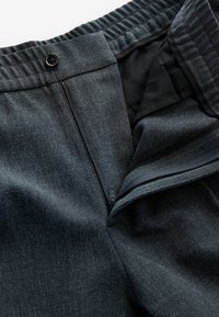 Mørkegrå bukser i stof med fleksibel linning, lynlås og knappelukning, der fremviser et tekstureret, vævet mønster.