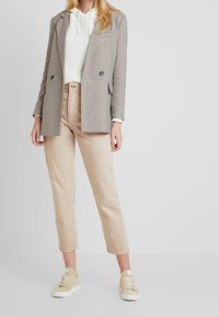 Tasiemkowy blazer w kratę z podwójnymi guzikami, zestawiony z białym, teksturowanym hoodie, jasnobeżowymi spodniami i beżowymi sneakersami.