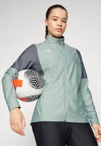 Svijetlozelena sportska jakna s patentnim zatvaračem i tamnosivim rukavima, uparena s crnim hlačama. Model drži bijelu nogometnu loptu s odvažnim uzorcima.