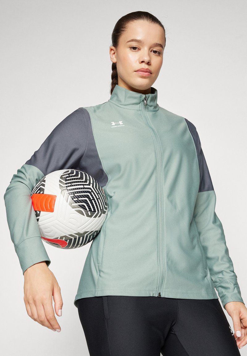 Giacca atletica verde chiaro con zip e maniche grigie scure, abbinata a pantaloni neri. Il modello tiene in mano un pallone da calcio bianco con motivi audaci.