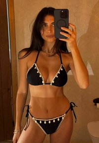 Czarny bikini z trójkątnym topem i regulowanymi wiązaniami, ozdobione białymi muszlami i fakturowanymi detalami. Neutralne tło.