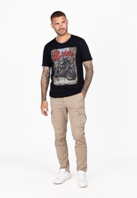 Camiseta gráfica negra con estampado de motocicleta, pantalones cargo beige con bolsillos laterales y zapatillas blancas. Ajuste casual y diseño simple.