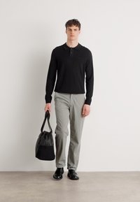 Sort langærmet polo-shirt, lysegrå bukser, sorte sko, holder en sort og grå tote-taske, står mod en neutral baggrund.