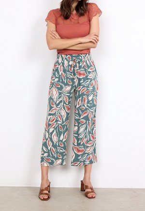 Groene wijde broek met bladmotief in rood en wit, voorzien van een elastische tailleband en een lichtgewicht stof. Bruine sandalen met bandjes.