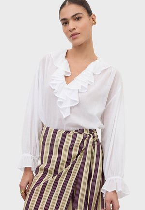 Femme portant un chemisier blanc avec un col et des poignets à volants superposés, assorti à une jupe portefeuille rayée verticale beige et violette.