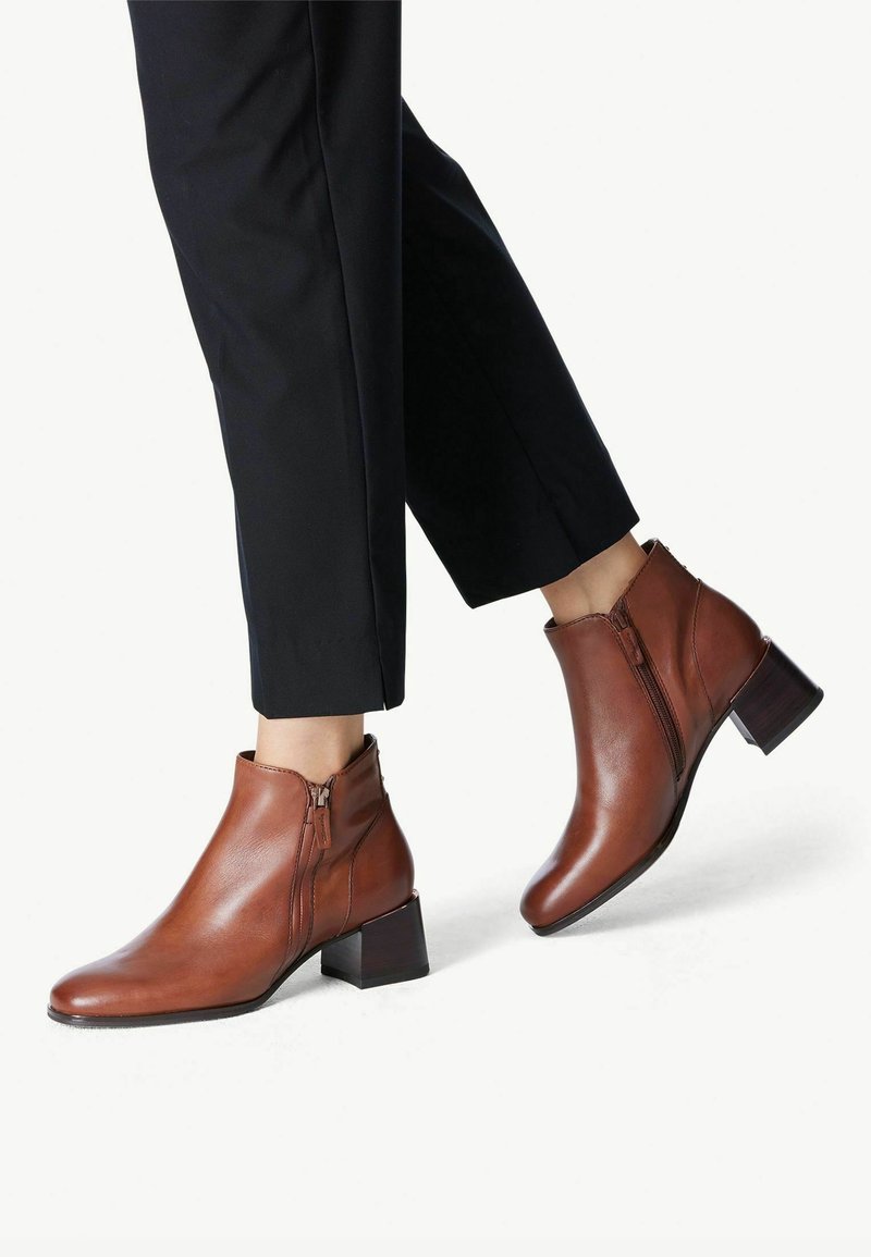 Bottines en cuir marron avec un petit talon carré, fermeture éclair latérale et un design élégant ; associées à un pantalon noir ajusté.