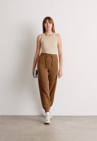 Top beige senza maniche e pantaloni jogger alti a vita alta, a sigaretta, marroni con cordoncino. La modella tiene una piccola borsa a tracolla con motivo. Scarpe da ginnastica bianche.