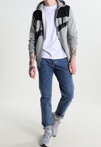 Muž oblečený v šedé a černé mikině, bílé tričko, modré džíny a bílé tenisky, stojící s viditelnými tetováními na obou předloktích.