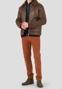 Uomo che indossa un giubbotto in shearling marrone, un maglione blu navy, una camicia a quadri, pantaloni color ruggine e stivali Chelsea marrone scuro, in piedi su uno sfondo grigio.