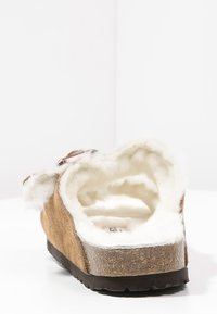 Zapatilla de ante marrón tipo slip-on con forro blanco esponjoso, suela de corcho texturizado y tacón redondeado. Presenta un interior suave y acogedor para mayor comodidad.