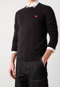 Pull en maille noire avec un col en V, arborant un petit logo rouge sur la poitrine. Superposé à une chemise blanche à col avec des boutons contrastants.