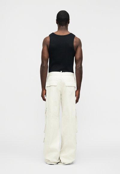 Άνδρας που βλέπει από πίσω, φοράει μαύρο αμάνικο τοπ και άνετο λευκό παντελόνι cargo με μεγάλες πίσω τσέπες, σε απλό φόντο.