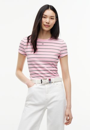 Jeune femme aux cheveux lisses et foncés portant un t-shirt rayé rose et un pantalon blanc avec une ceinture blanche sur un fond uni.
