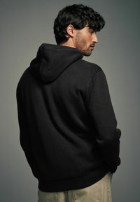 Mustmust särk, millel on tekstureeritud kangas, soonilised varrukad ja äär. Kandja pööratud seljaga, näidates kapuutsi ja külgprofiili neutraalse tausta taustal.