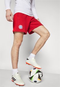 Shorts de sport rouges avec le logo du FC Bayern Munich, portés avec un haut ajusté à manches longues blanc. Chaussures de football blanches sur un ballon multicolore.