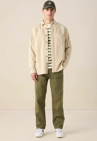 Beige corduroy overhemd, gestreepte groen-witte t-shirt eronder, olijfgroene broek en witte sneakers met rode accenten.