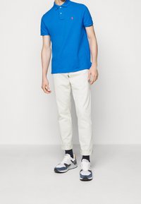 Niebieska koszulka polo z kołnierzykiem i trzema guzikami, kremowe taperowane spodnie oraz białe sneakersy z niebieskimi i czarnymi akcentami.