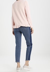 Top lungo a maniche con rouches di colore rosa chiaro, abbinato a jeans skinny blu medio con un pannello laterale blu scuro e orli sfrangiati. Sneakers bianche.