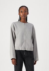 Grå, plysj zip-jakke med rund hals, senkede skuldre og lange ermer, kombinert med mørke jeans. Teksturert stoff synlig.