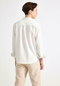 Kremowa koszula z długim rękawem, z kołnierzykiem i zapięciem na guziki przy mankietach, z luźnym krojem i subtelną teksturą. Noszona z jasnobeżowymi spodniami.