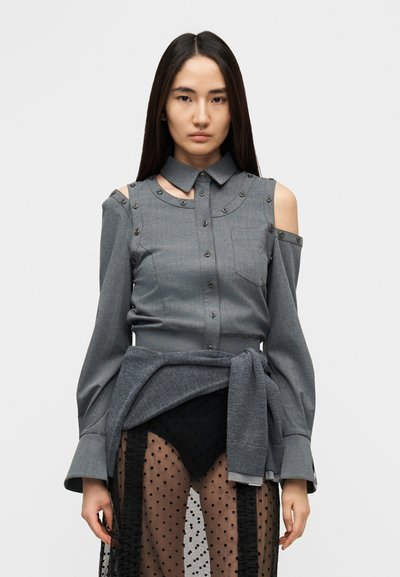Chemise grise boutonnée avec découpes aux épaules, boutons décoratifs, col structuré et manches longues. Superposée sur un tissu noir transparent à pois.