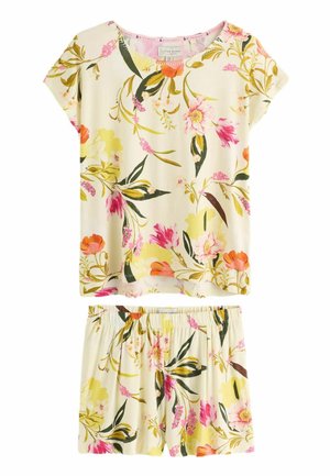 Frau steht drinnen und trägt ein geblümtes Kurzarmkleid, mit langen dunklen Haaren, Bett mit Kissen und Decken im Hintergrund.