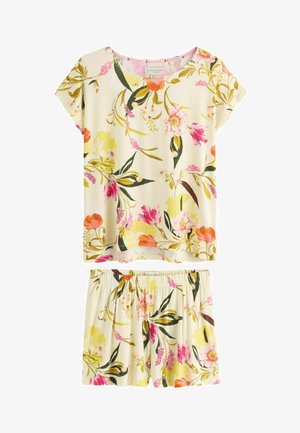 Žena stojaca v interiéri oblečená vo kvetinových šatách s krátkym rukávom, s dlhými tmavými vlasmi, posteľ s vankúšmi a prikrývkami v pozadí.