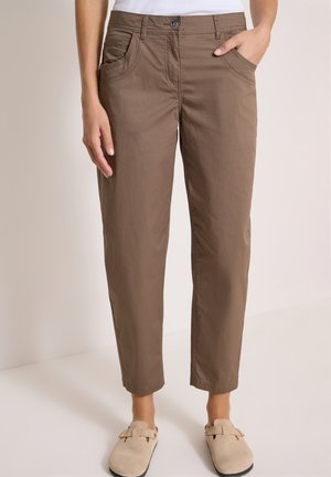 Personne portant un pantalon marron droit avec des poches avant et des chaussures beiges à enfiler, debout devant un fond clair neutre.