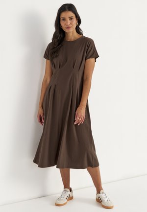 Vrouw draagt een bruine midi-jurk met korte mouwen en sneakers, staat tegen een witte muur, handen ontspannen langs het lichaam.