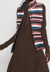 Viskoitu takki, jossa on monivärisiä vaakaraitoja (vaaleanpunainen, sininen, musta) ruskean ribbimekon päällä. Varustettu vetoketjulla ja korkealla kauluksessa.