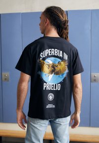 Črna bombažna majica z grafiko orla na modrem krogu, z besedilom "SUPERBIA IN PROELIO" zgoraj in logotipom spodaj.