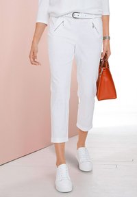 Weiße, verkürzte Hosen mit umgeschlagenen Säumen, vorderen Reißverschlusstaschen und einem weißen Gürtel. Kombiniert mit weißen Sneakern und einer orangefarbenen Handtasche.