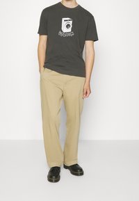 T-shirt grafica grigia con design di una lavatrice, abbinata a pantaloni beige a gamba dritta e scarpe nere con cuciture gialle.