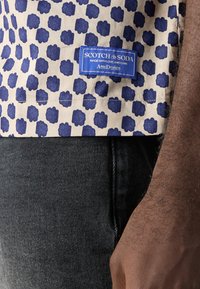 Camisa de manga corta con fondo beige y patrón floral en azul marino, con una etiqueta azul que dice "Scotch & Soda" en el dobladillo.