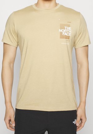 Homme portant un t-shirt beige North Face avec logo et texte sur le côté gauche de la poitrine et un pantalon noir avec un petit logo blanc sur la cuisse gauche.