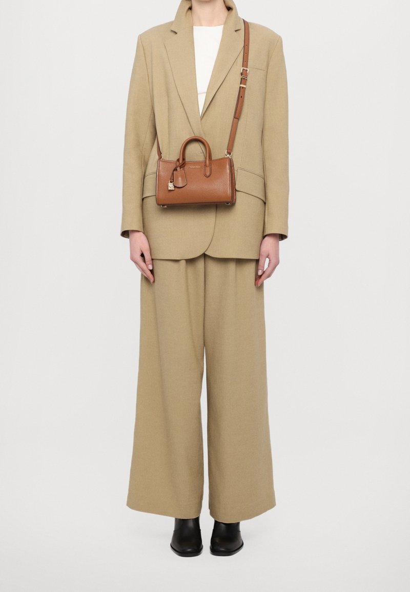 Personne portant un costume beige oversize, un haut blanc, des bottes noires et portant un petit sac à main en cuir marron avec une bandoulière.