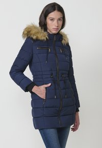 Giacca a vento blu navy con cappuccio in pelliccia sintetica, tasche con zip e coulisse regolabili in vita; presenta un design trapuntato e dettagli in hardware dorato.