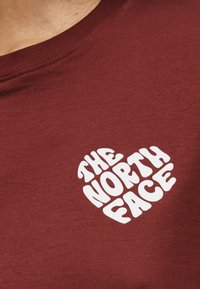 Μπορντό πουκάμισο με λευκό κείμενο σε σχήμα καρδιάς που γράφει "THE NORTH FACE" στο πάνω μέρος του στήθους κοντά στη λαιμόκοψη.