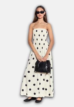 Weißes Maxikleid mit schwarzen Polka Dots, tailliertem Oberteil, gerafften Details und fließendem Rock. Kombiniert mit einer kleinen schwarzen Fransentasche.