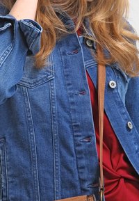 Džínsová bunda s klasickým střihem, modrá barva, přední kapsy, stříbrné kování a knoflíkové zapínání, přeložená přes červený top. Lehké textury.